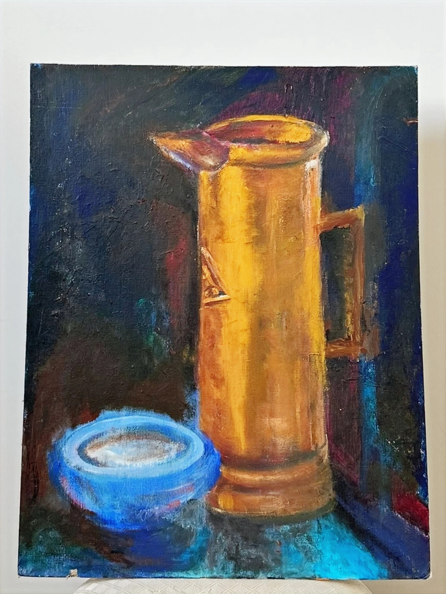 A tall golden copper pitcher beside a small blue bowl on a dark blue surface. Still Life With Copper Pitcher, by Evelyn Altman.