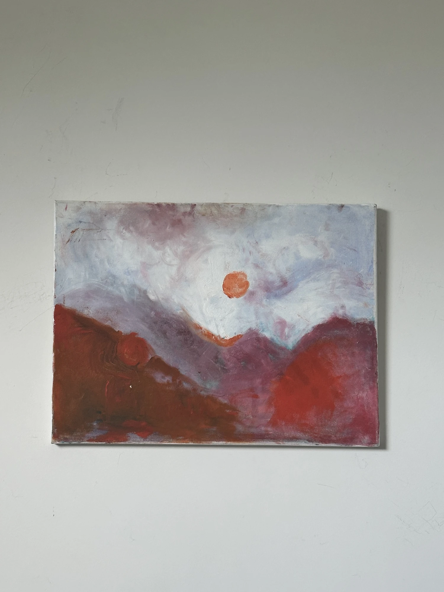 A small orange sun in a pale misty sky above overlapping deep red mountain forms. The Sun Blessing + Devouring, by Evelyn Altman.
