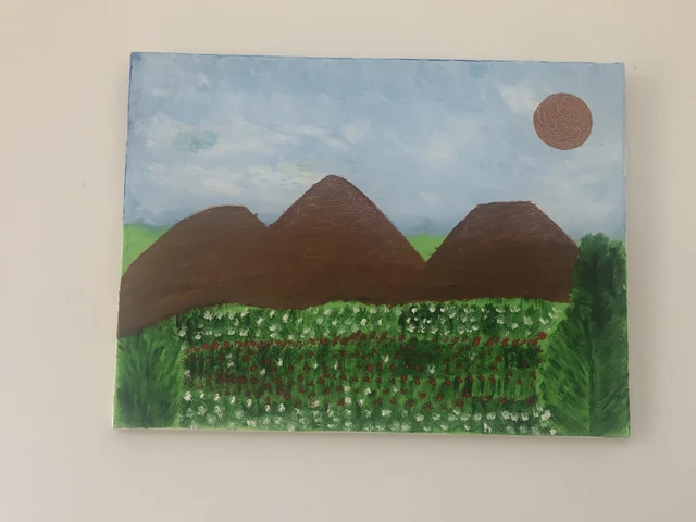 Three brown mountain peaks above a green meadow of small white flowers, beneath a pale blue sky and rust-colored sun. Green Meadow at The Mountain, by Laida Altman.